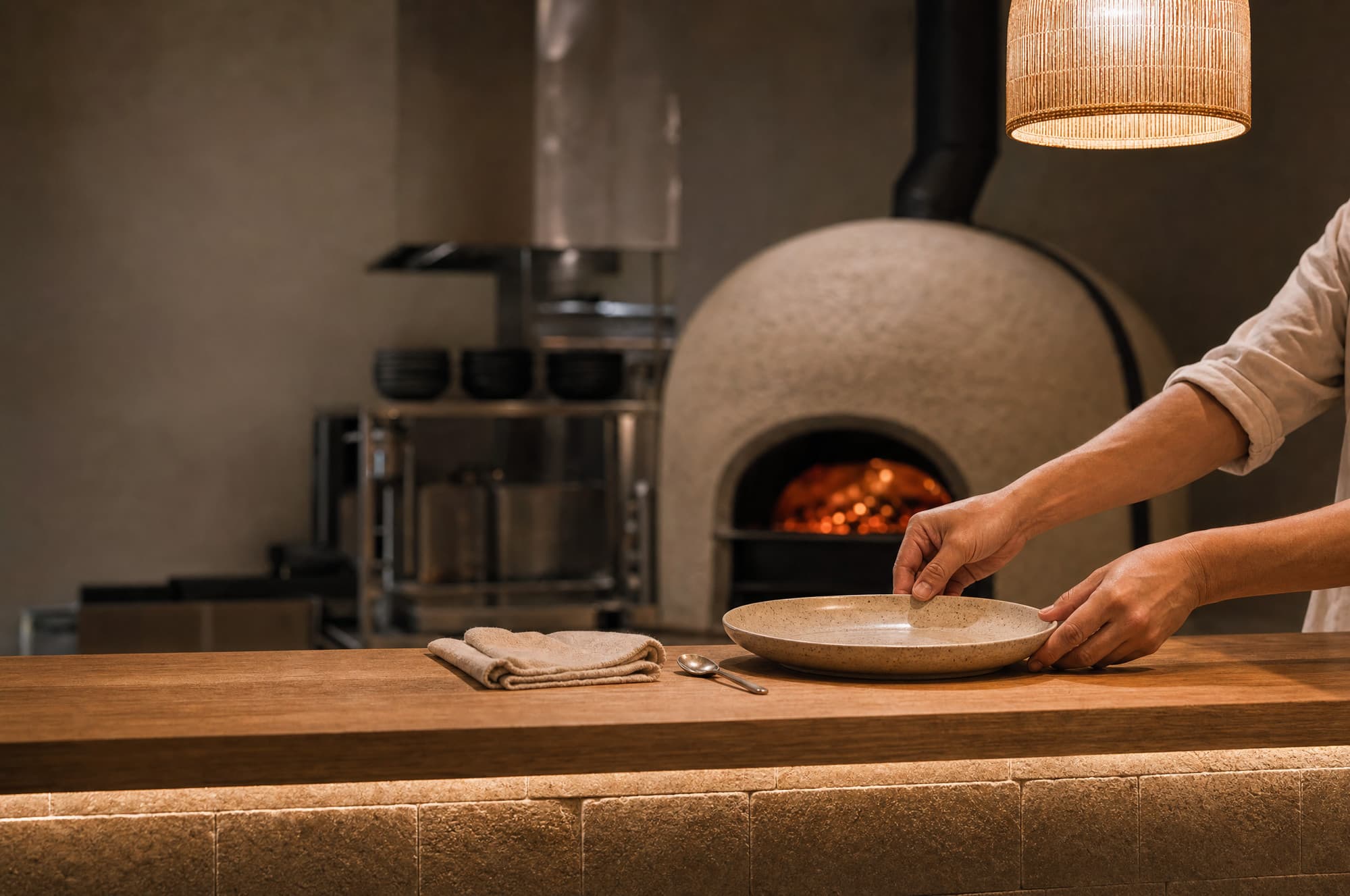 A server plating a dish at the pass, wood-fired oven behind the counter